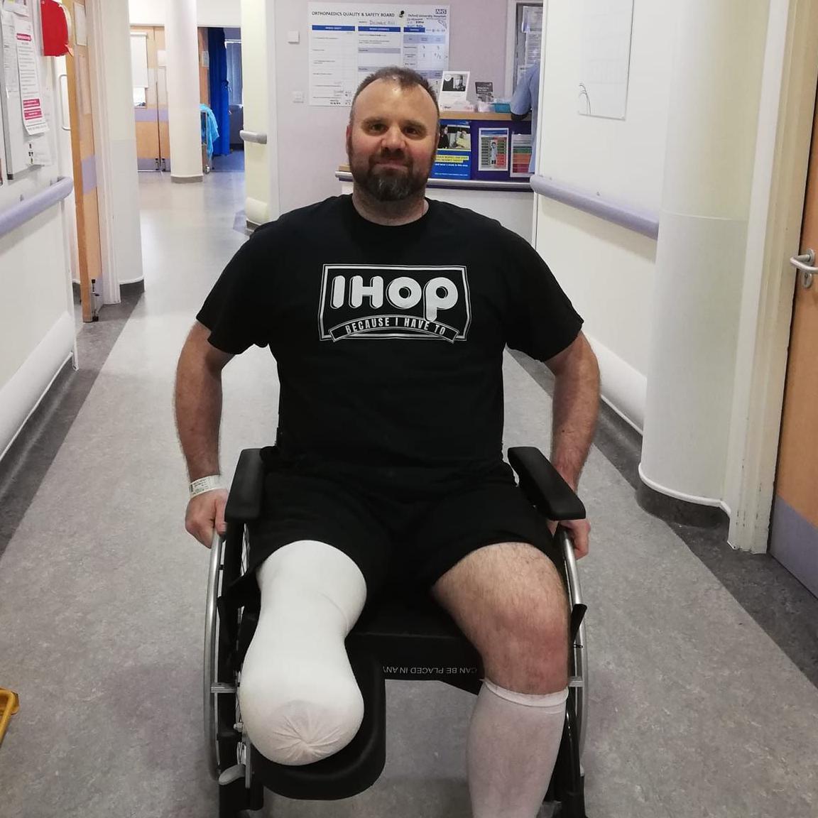 Mark Willerton in a wheelchair in a hospital corridor. He is wearing a black T-shirt with an Ihop logo, and black shorts. His residual limb post-amputation is bandaged. He is smiling.