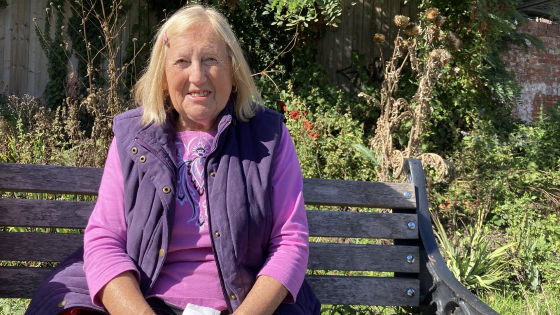 A woman with blonde shoulder length hair, wearing a purple vest over a pink top sits on a wooden bench in a sunlit garden, surrounded by green plants and trees, with some dried vegetation in the background.