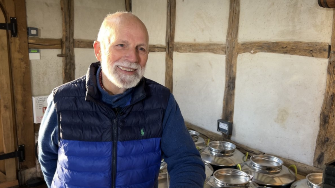 Charles Martell has a grey beard and is wearing a blue jacket in front of some barrels 