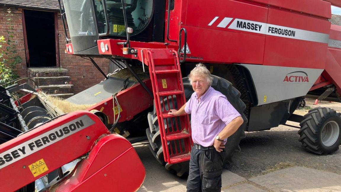 A man with a purple shirt and dark trousers is standing next to a farming vehicle, which is mainly red.