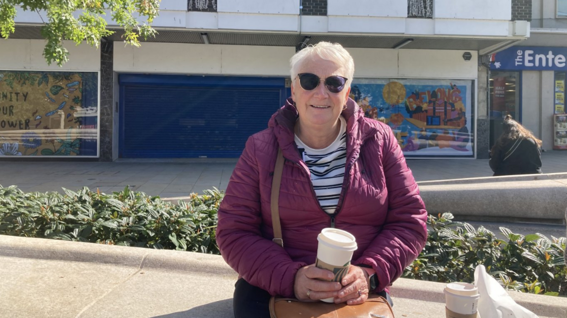 A woman sits on a stone bench in a sunlit urban setting, holding a coffee cup and wearing a maroon jacket, striped shirt, and sunglasses. A brown handbag rests on her lap, with two additional coffee cups and a white paper bag placed beside her. Trees and greenery frame the scene.
