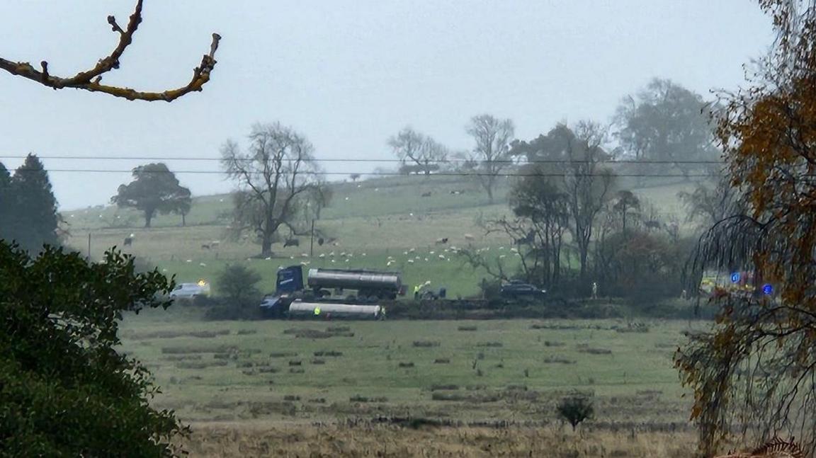 A distant shot of a two tankers, one on its side in a field and the other on the road. People in high-vis jackets can be seen as well as emergency vehicles. It's a rural scene with sheep visible in the field behind.