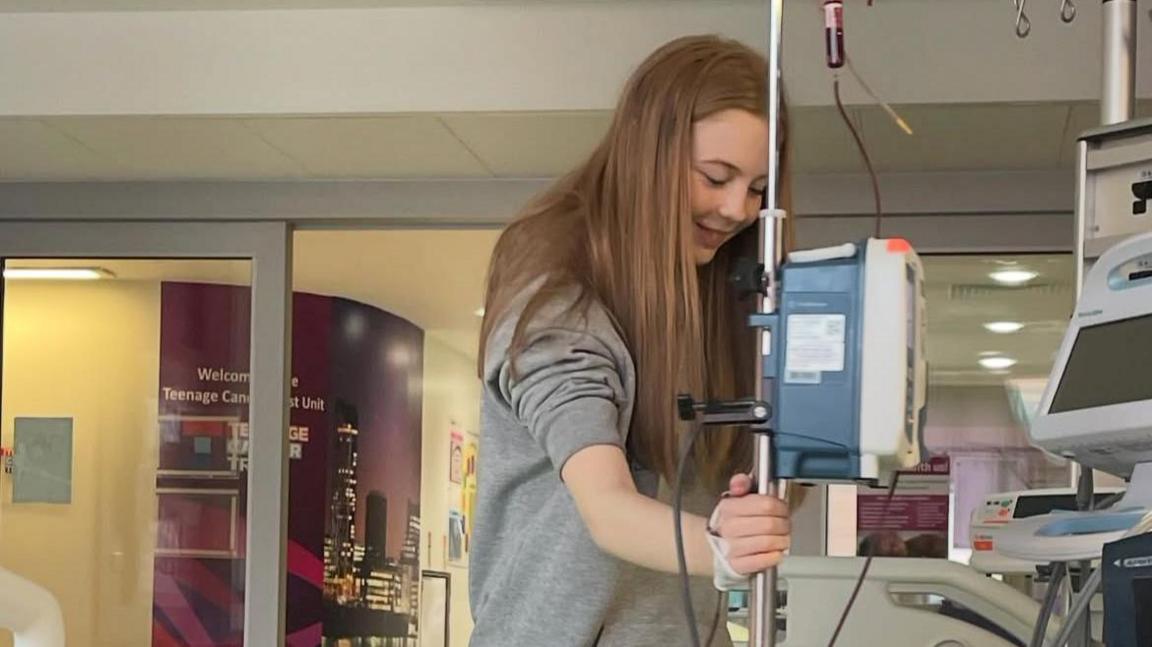 Maisie has long red hair and is wearing a grey jumper. She is in hospital while she has a blood transfusion.
