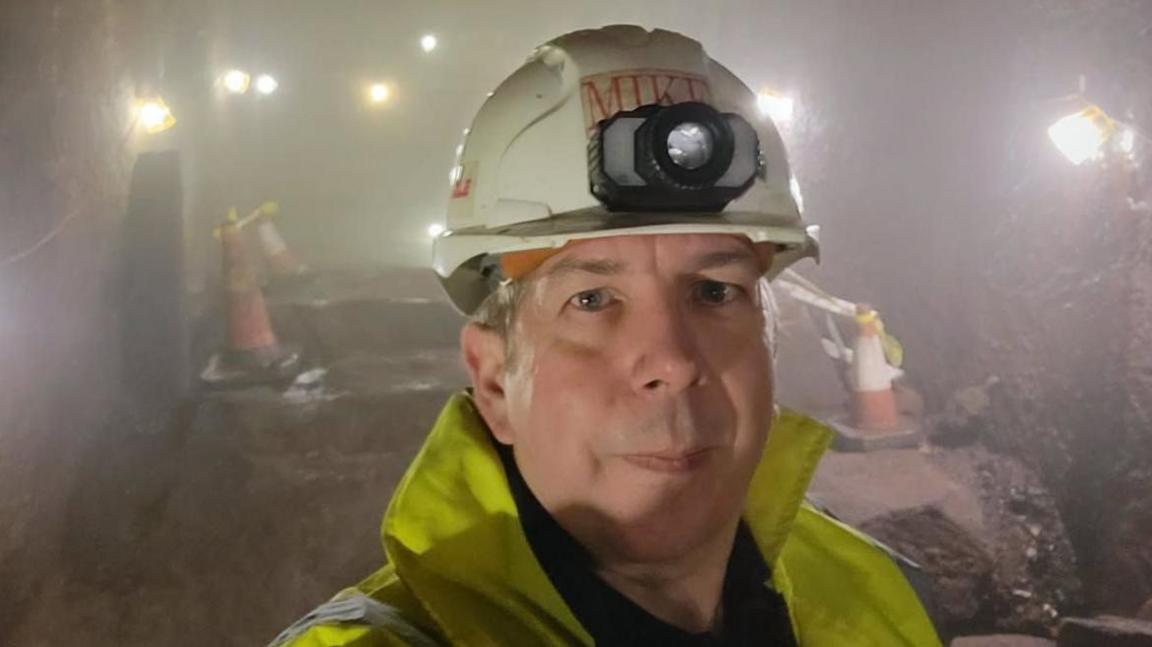 Mike Rogers, who is wearing a white hard hat with the name 'Mike' across the front, and a yellow high-visibility jacket, is pictured inside a tunnel