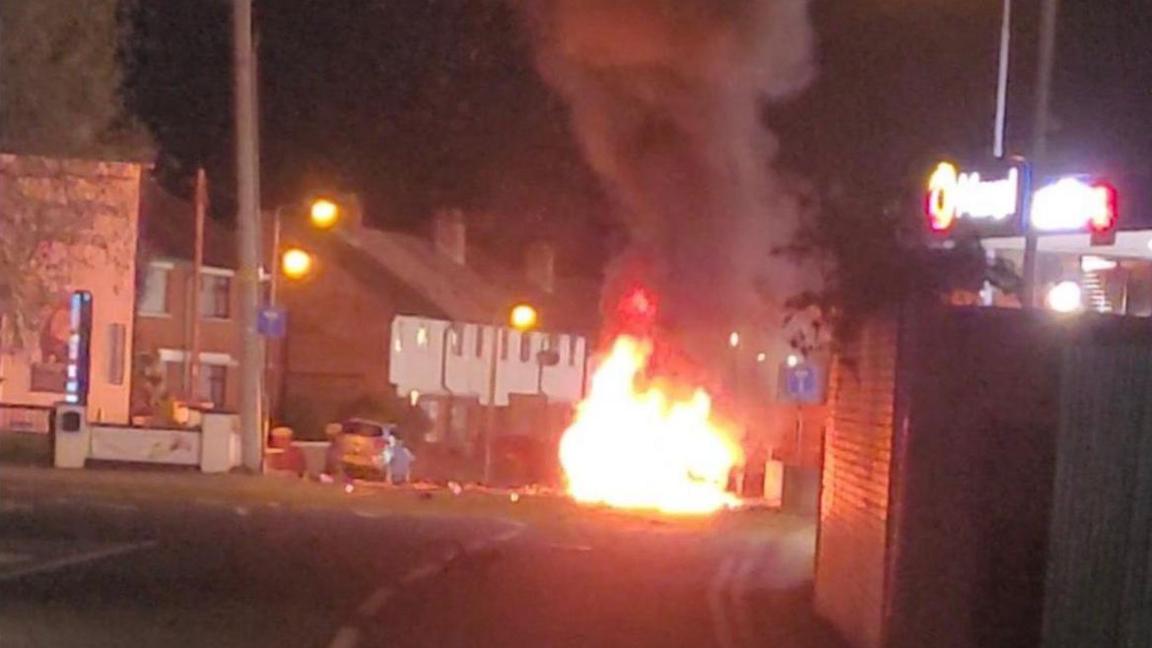 At night on a road, a car is on fire near a building. Bright flames and thick black smoke rise into the air. Streetlights and nearby houses can be seen in the background.