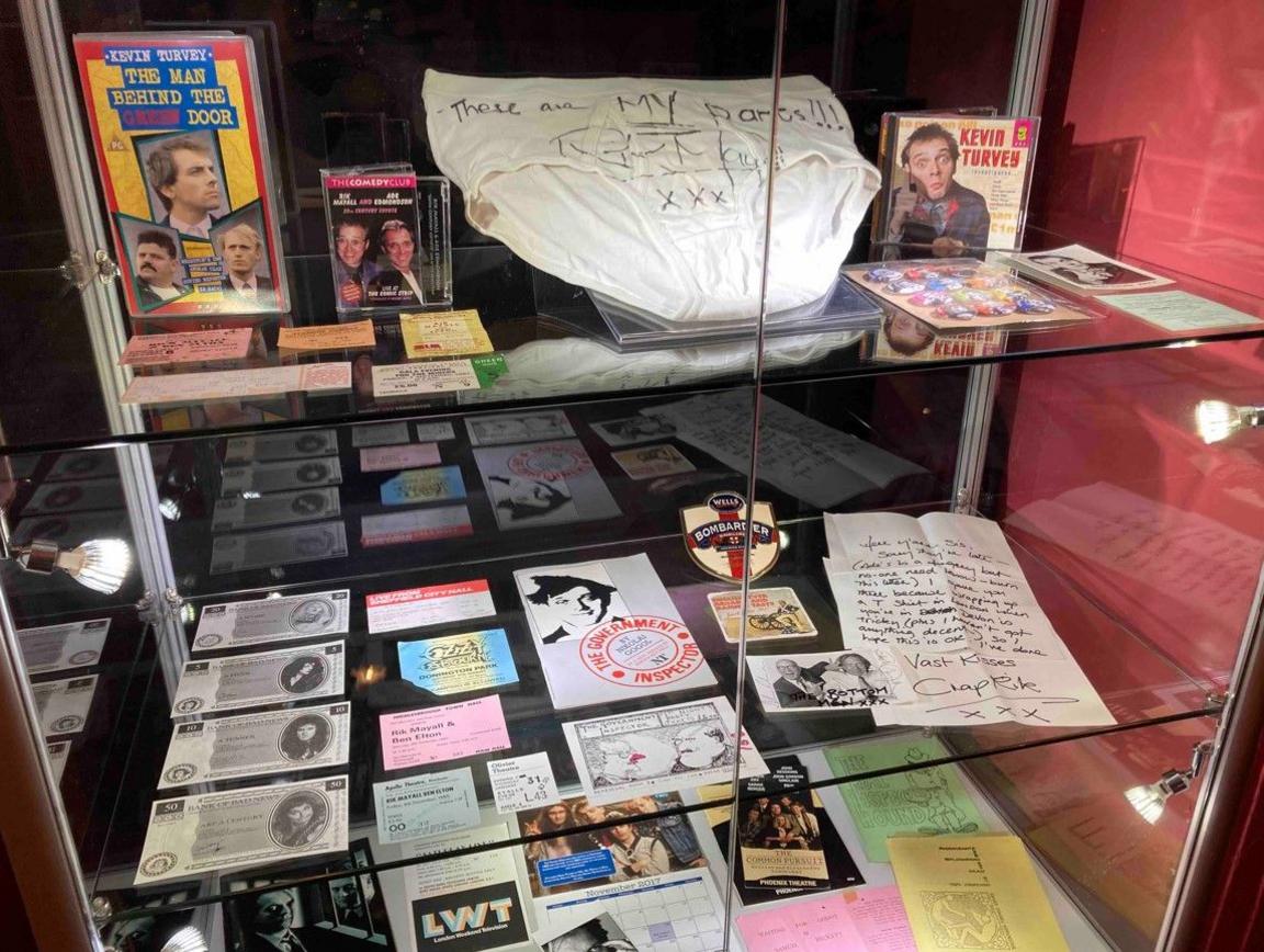 Memorabilia is visible on glass shelves. The top shelf includes recordings of Mayall, the next shelf features tickets and a letter and the bottom area includes photos.