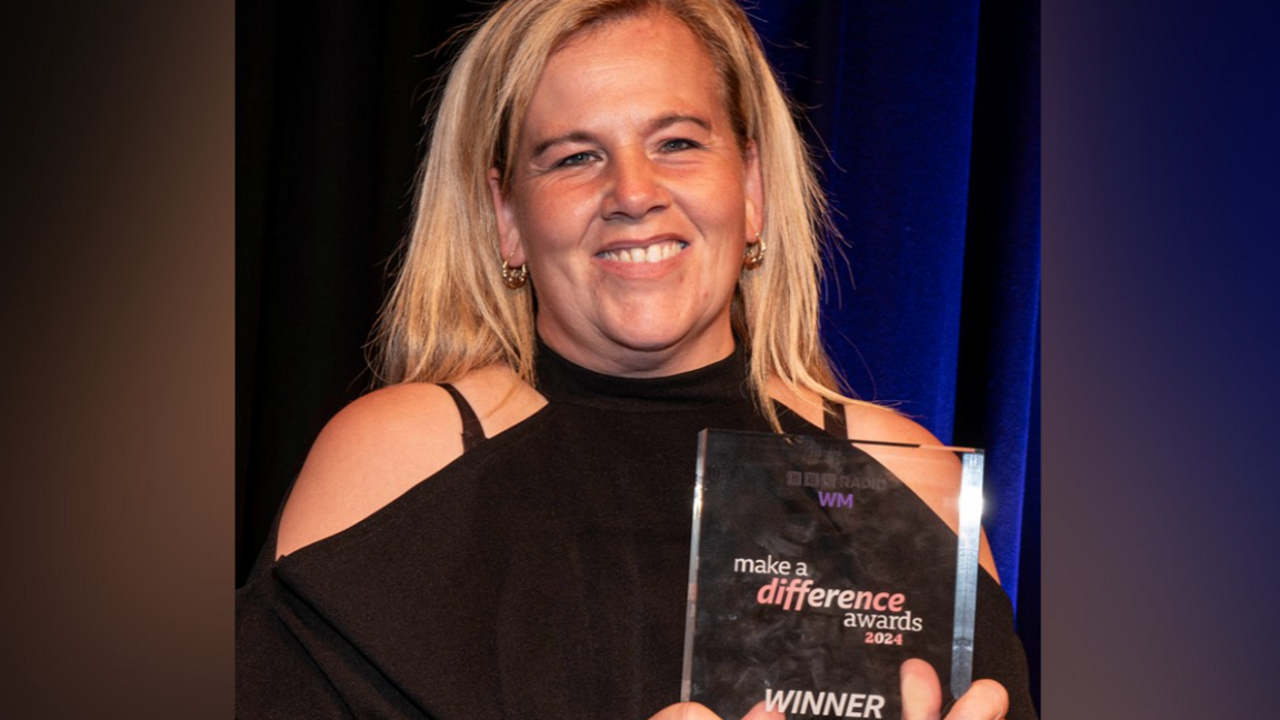 Sam Steele is wearing a black top with the shoulders cut out and has blonde hair and is smiling in the photo while holding her award