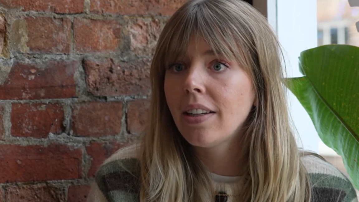Sam Sheldon sitting in front of a brick wall with the edge of a large pot plant beside her