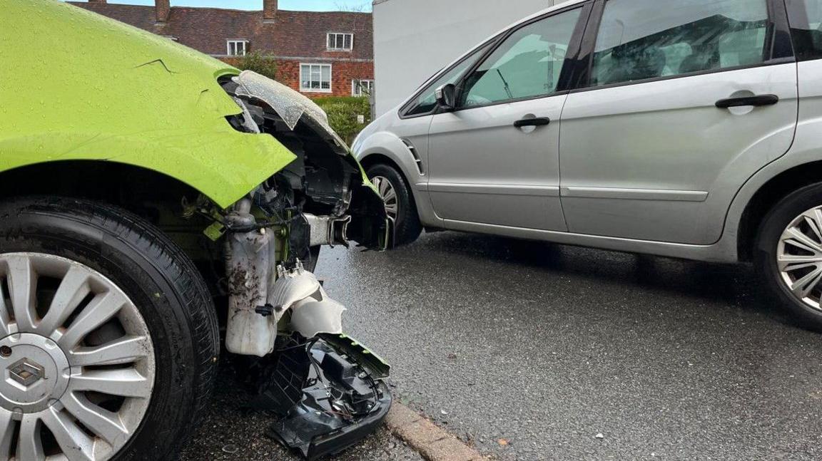A close up image of significant damage to the front bumper of a neon green Renault car which is parked very close to a tight section of road being used by cars and vans with houses in the background.
