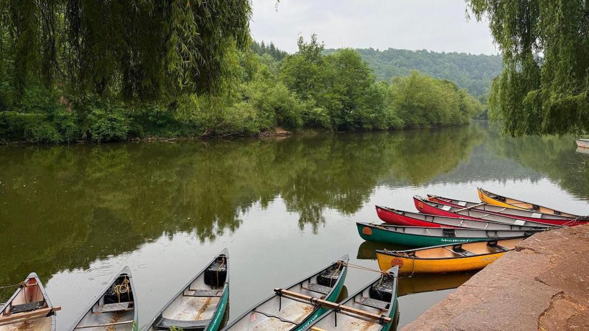 A series of boats tied up on a lake with trees on the far bank, reflected in the water. The boats are different colours, green, yellow and red.