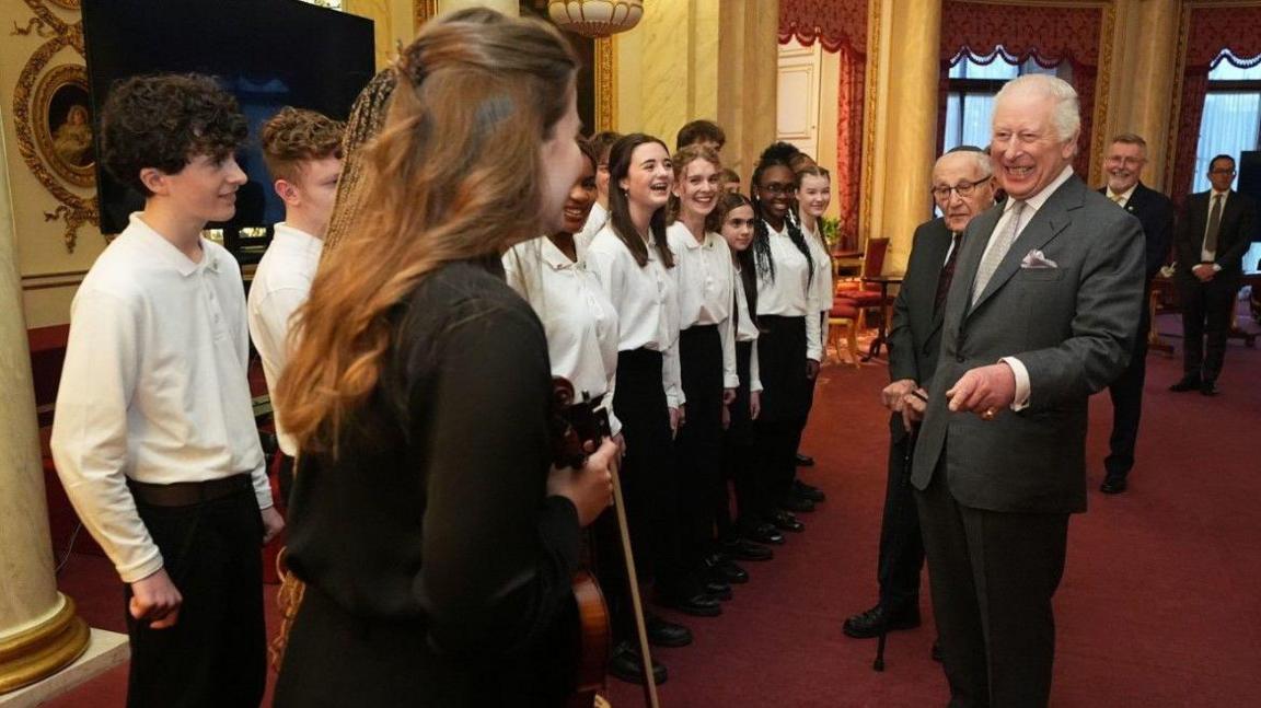 More than 10 students in white tops are visible on the left and centre of the photo. King Charles is on the right and wearing a grey suit, and three other men are in the background.