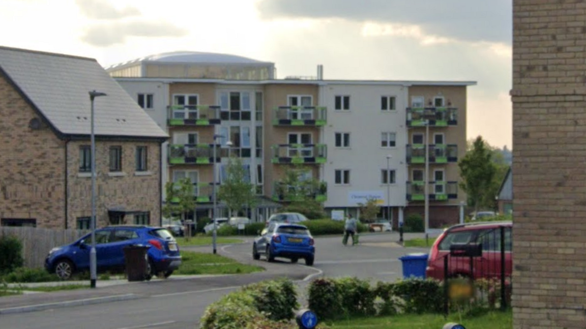 Clement House, a four-storey block of flats on a new-build estate. There are cars parked outside. In the foreground are houses.