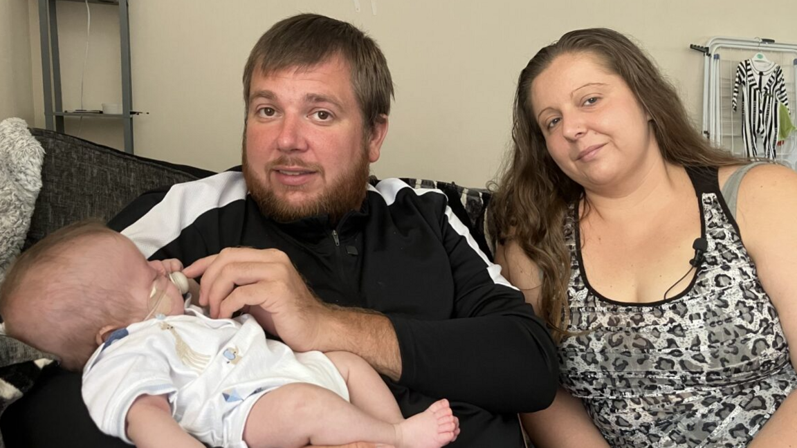 A man and a woman sitting on a sofa. The man is holding a baby. The couple are both looking into the camera.