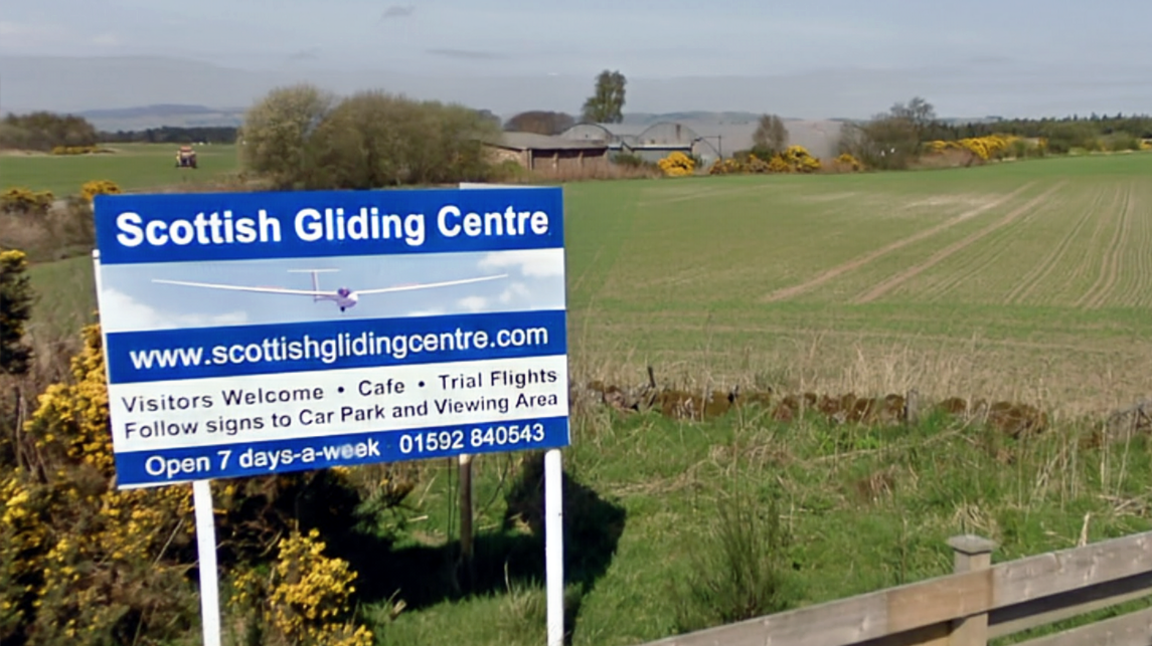 A rural road runs alongside open fields under a clear blue sky, with a sign for the Scottish Gliding Centre visible near a wooden fence.
