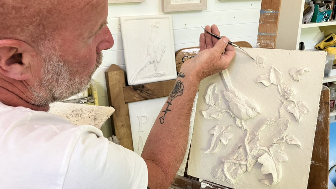 Ian Warren, with a close-cropped greying beard, shaved head and wearing a white t-shirt, works on a piece of art with white plaster on a canvas. It depicts a bird sitting on a branch, surrounded by leaves.