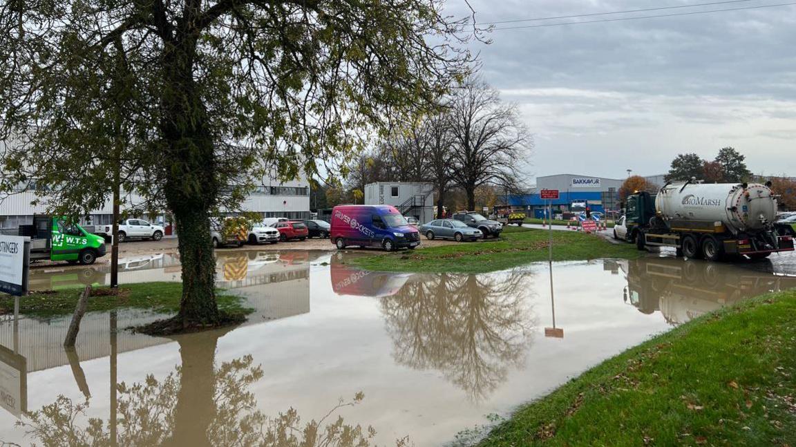 A car park flooded with brown murky water with cars dotted about and industrial buildings in the distance.