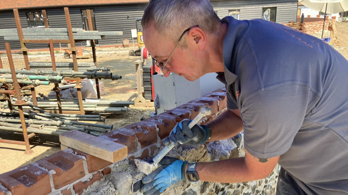 Mark Clifton, who has short grey hair and glasses and is wearing a grey polo shirt and blue work gloves, holds a flint stone in place on a partially-constructed wall 