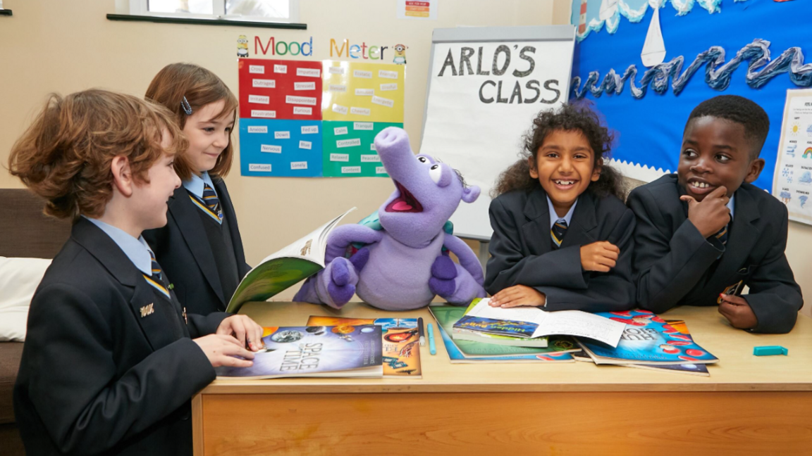 Four school pupils, all wearing dark coloured blazers, light blue shirts and ties, sit round a table smiling and looking at Arlo the Armadillo, a lilac cartoonish armadillo puppet. In the background is a whiteboard with "Arlo's Class" written on it.