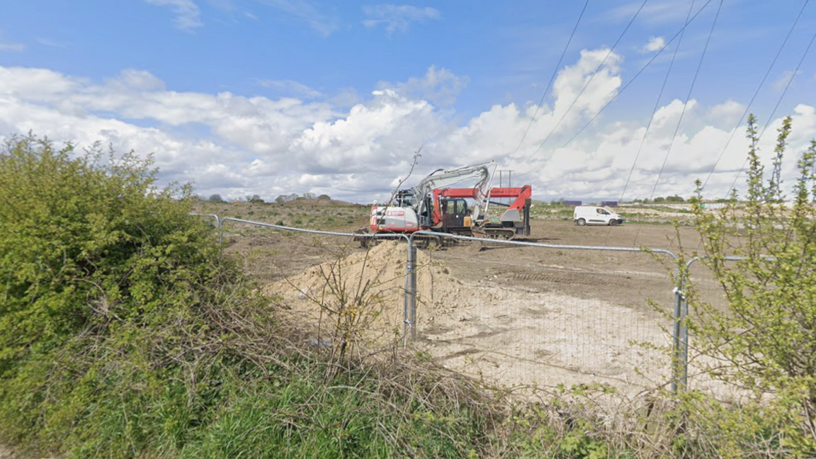 A picture of one of the fields where the solar farm will be built. A metal fence has been erected around the field next to a row of hedges. A digger is parked in the field near to a white van and a pile of earth.