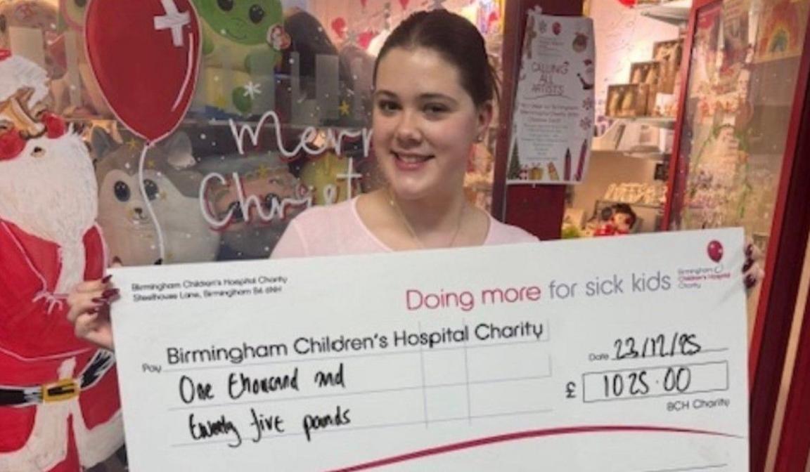 Ms Giorgio-Jones holding up a large cheque to Birmingham Children's Hospital  Charity for £1,025. She is in front of Christmas images and words in a window.