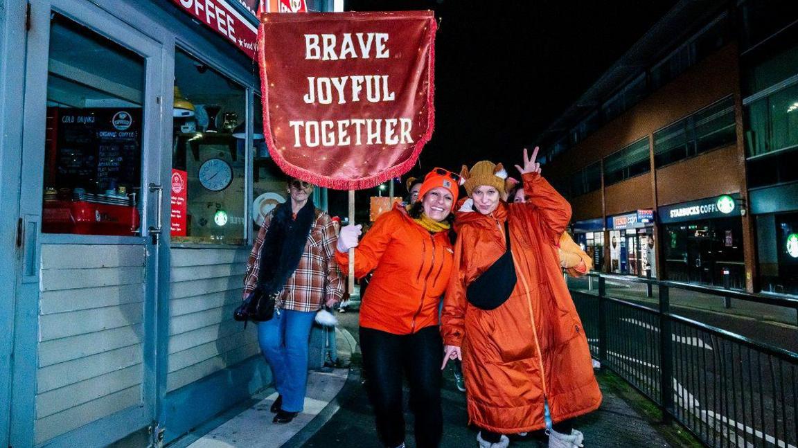 Two women wearing orange coats pose on the pavement next to a road. One of them is holding up a sign saying "brave, joyful, together".