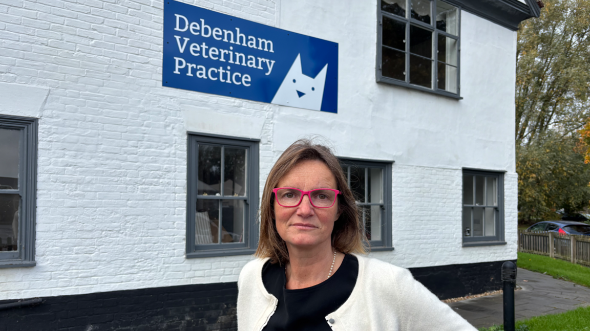 A head and shoulders image of Jade Martin from Debenham Veterinary Practice. She is wearing a white cardigan over a black top and pink glasses. She is looking into the camera while standing in front of the vet practice's white building.