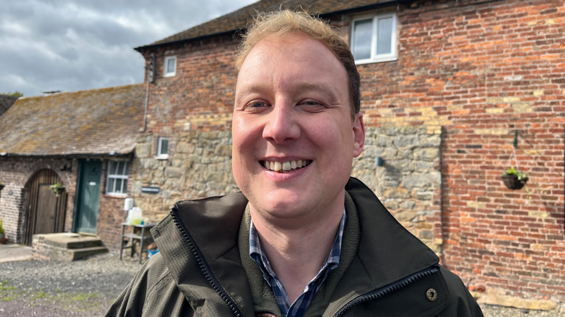 A man in a waxed jacked smiling at the camera and standing in front of farm buildings