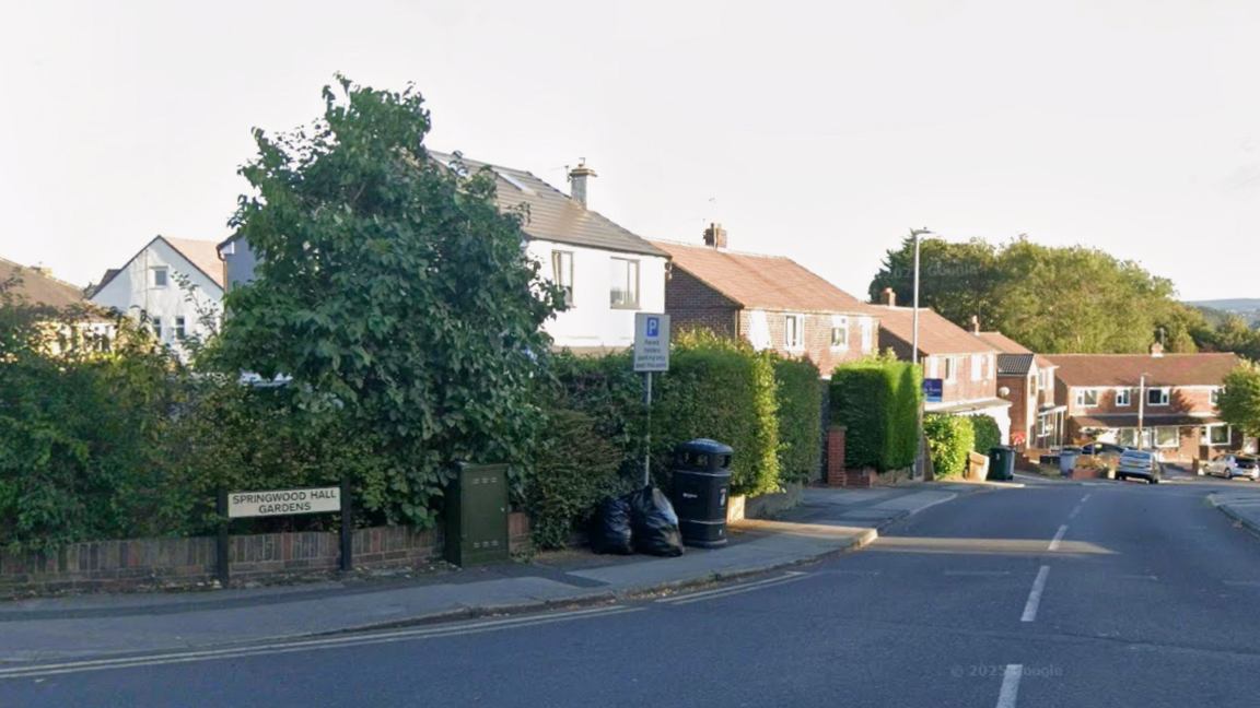 Residential street with semi-detached houses featuring red-tiled roofs and white walls. A large leafy tree and trimmed hedges line the pavement. There is a street sign reading ‘Springwood Hall Gardens’ on the left, along with a parking restriction sign and two black rubbish bags near a bin.