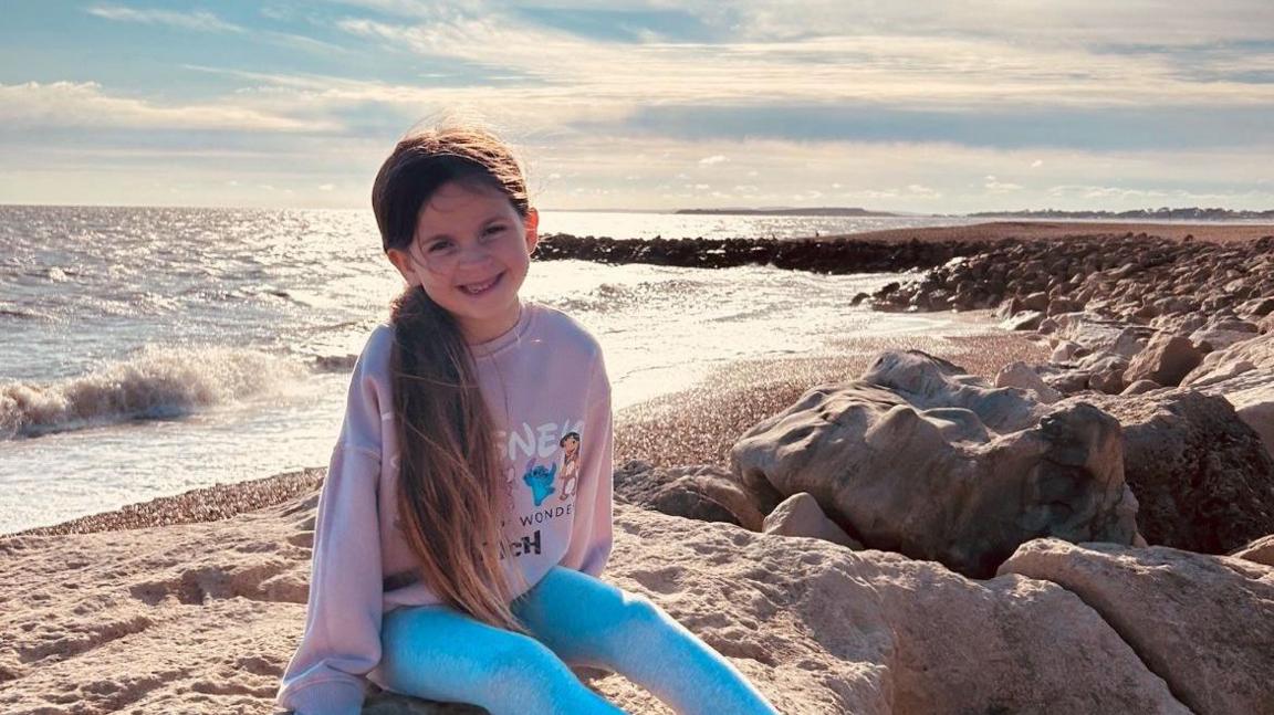 A young girl wearing grey leggings and a pink Disney sweatshirt. She sat on some rocks with the sea in the background. 