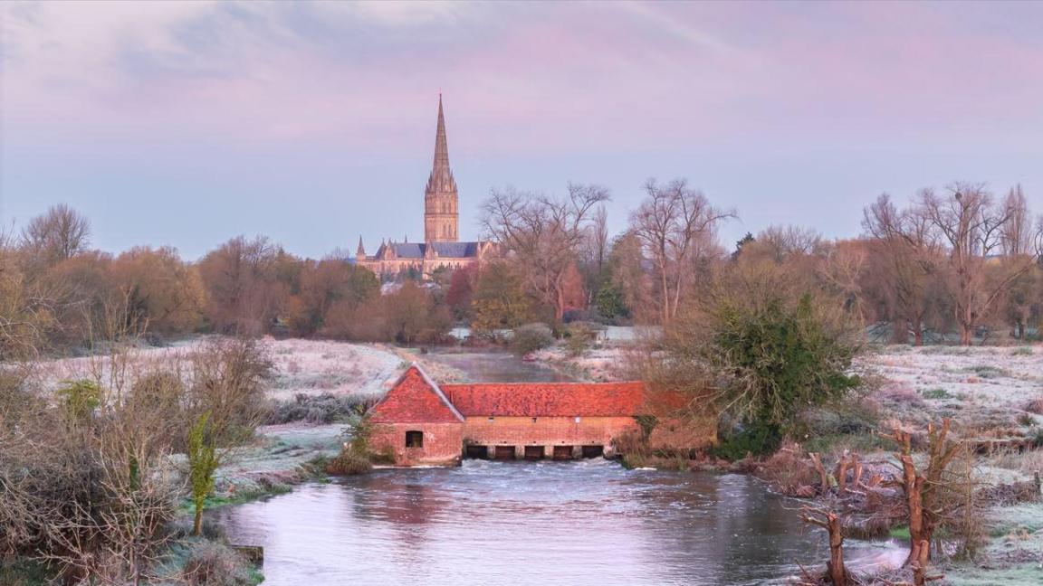 A calming scene overlooking the River Avon towards Salisbury Cathedral in the distance. The sky is pink, light blue and purple, and the surrounding grass is covered with sparkling frost. In the middle of the river there is a long brick building with several tunnels running underneath it. 