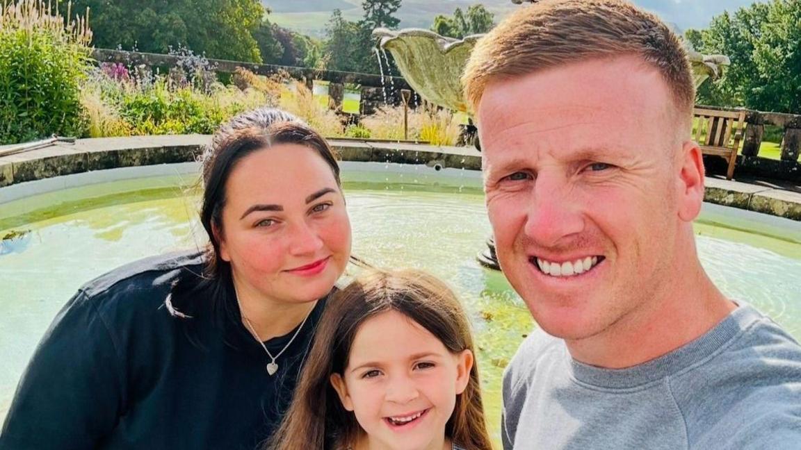 A selfie taken of the Smith family, they are next to a water fountain. On the left is a woman with brown hair wearing a silver heart necklace and a black t-shirt. In the middle a young girl with brown hair, on the right a man with ginger hair and a grey jumper. 