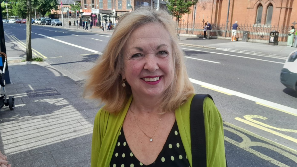 A woman with blonde shoulder length hair smiles into the camera. She's standing on a footpath wearing a green cardigan and blacky spotty top.