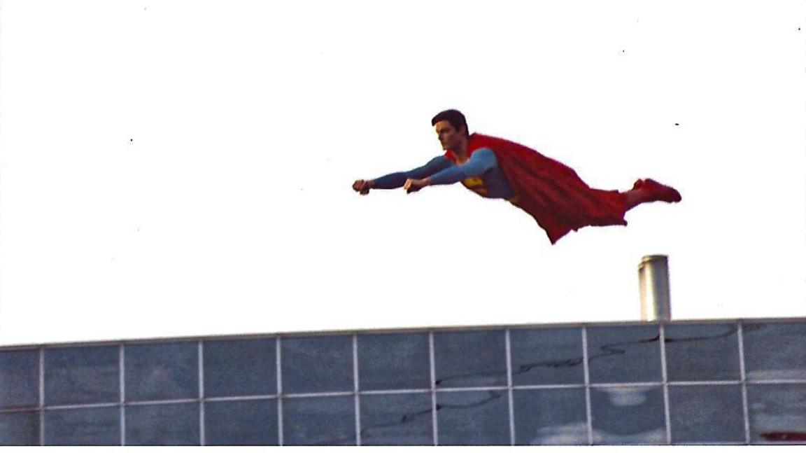 Christopher Reeve wearing a Superman costume with his hands out stretched in front of him as if he is flying, suspended over a modern building with a flat roof and a short silver chimney.