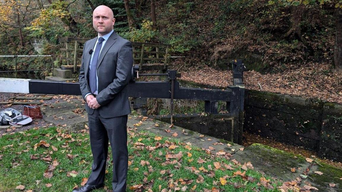 A picture of Det Ch Insp James Entwistle who is wearing a dark gre coloured suit and tie and is stood next to a river.