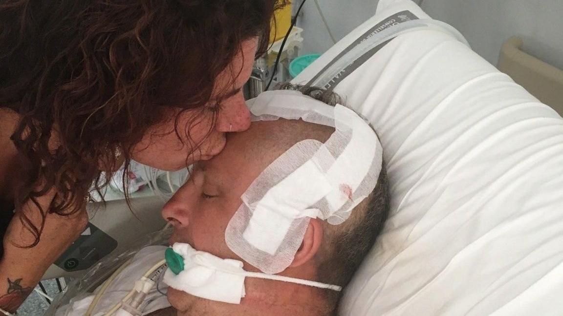 A young woman kissed the forehead of a man in a hospital bed. He has bandages all over his head and tubes in his mouth and nose. 