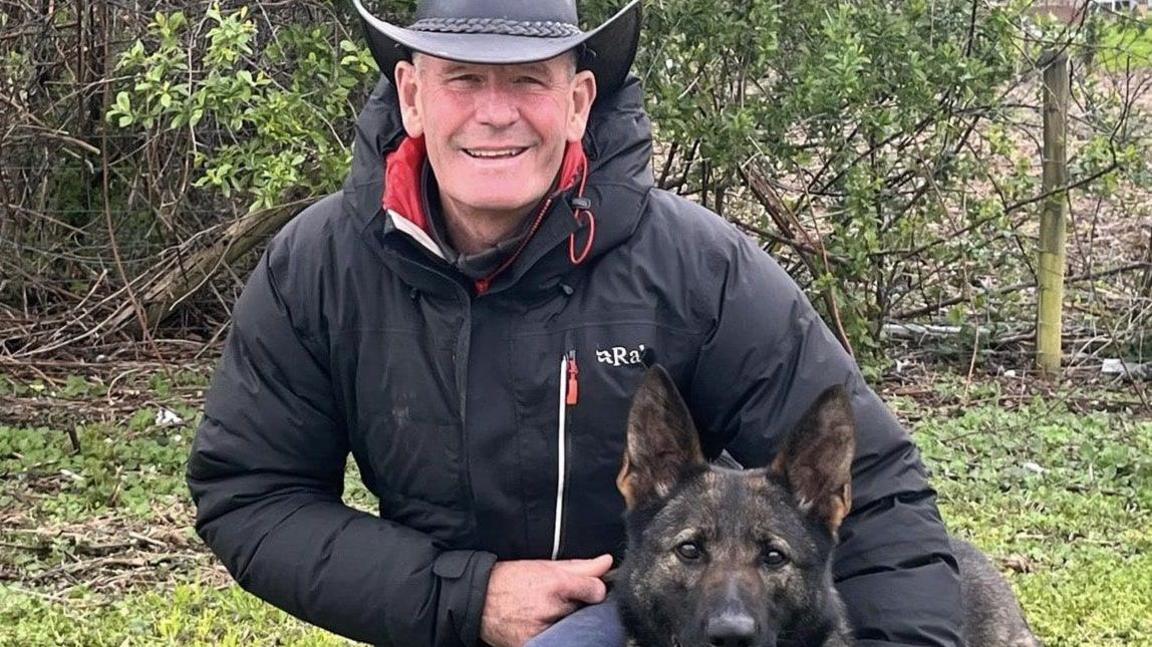 Martin Winfield wearing a black hat and a black jacket. He is smiling with a black dog just his right. The pair are in a rural, wooded area with bushes seen in the background.