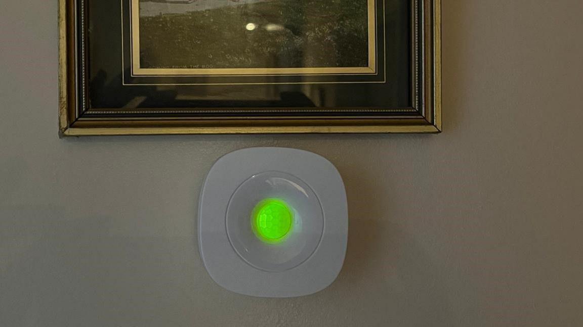 A white plastic sensor is on the wall with a green light in its centre. It sits on the wall below a black and gold picture frame.