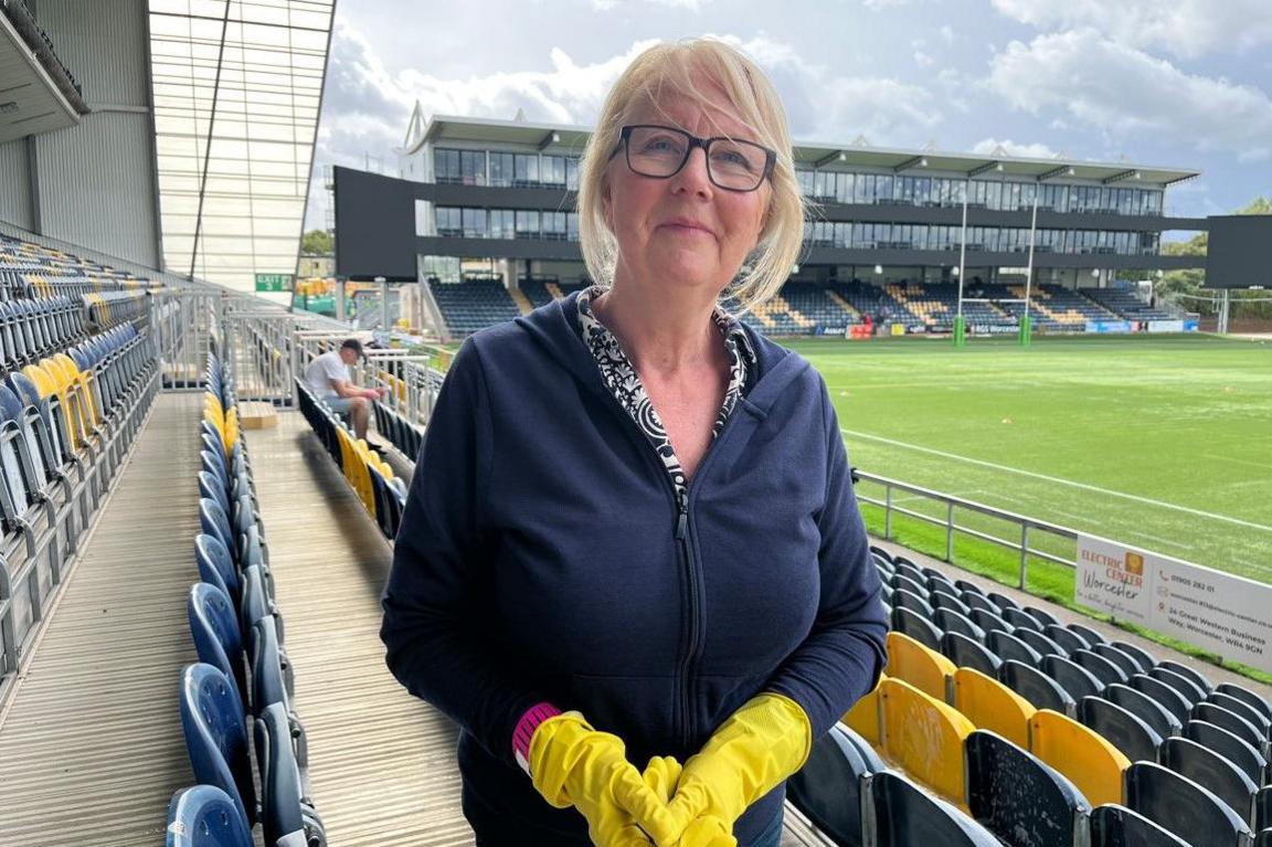A woman with blonde hair and wearing a blue top is wearing yellow gloves. Seating is directly behind her and another stadium stand is in the background.