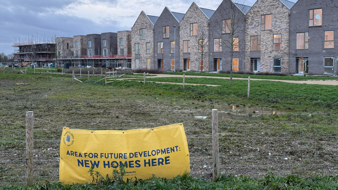 A muddy field with a fence around it. On the fence is a yellow sign reading 'area for future development - new homes here'. Behind the field is a row of new build home, some are still under construction.
