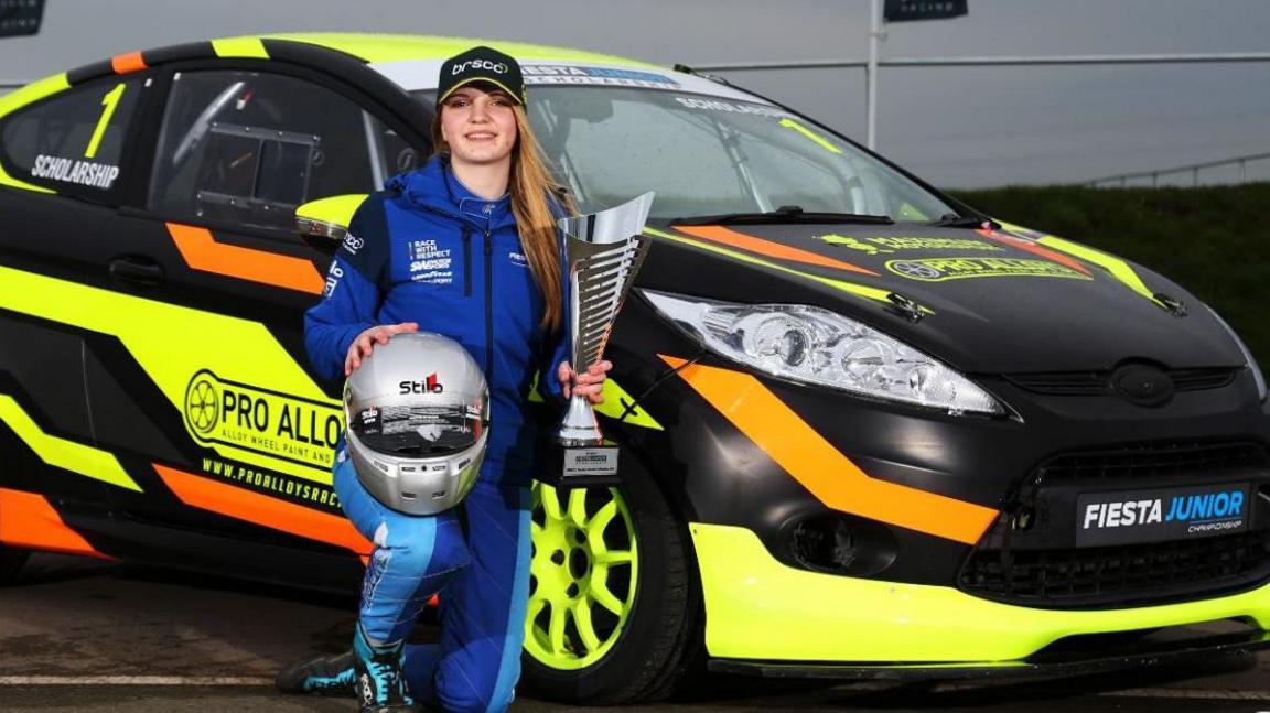 Daniella Sutton is kneeling next to a Ford Fiesta car which has branding of Fiesta Junior. She is wearing a blue racing suit and a baseball cap and is holding a racing helmet and a trophy.