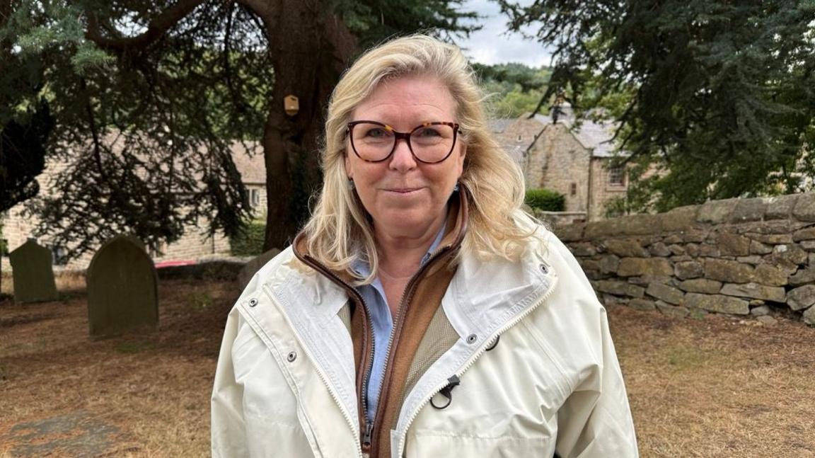 A person wearing prescription glasses and a light-coloured jacket standing outdoors beneath a large Atlas Cedar tree. A couple of old gravestones are visible in the background, along with a stone wall.