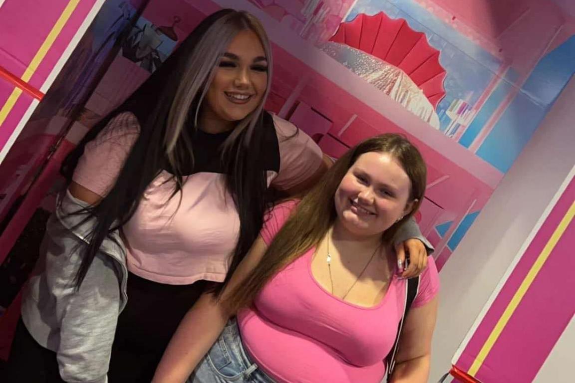 Two teenagers, Megan and Lily Nichol, are stood inside a human-sized pink cardboard Barbie box. They are both wearing pink tops and are smiling at the camera