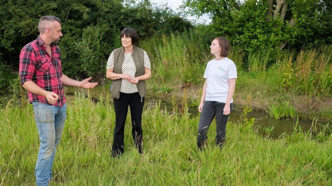 Will Shemilt with short light brown hair wearing a red and black shirt with blue jeans. He has both hands outstretched while explaining something. Kim Hemmings has medium-length dark hair and is wearing a white T-shirt, green gilet and dark trousers. Hannah Worker has long dark hair, a light-coloured T-shirt and dark trousers. Both of the women are looking at Will. They are standing on grass with a pond and trees behind them.