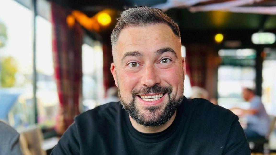 A man with short hair and a short, dark beard, wearing a black jumper. 