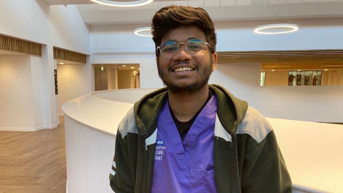 Badusha Mohammed, with dark hair, is wearing a green top, with purple NHS clothing underneath it. He is wearing glasses and smiling at the camera.