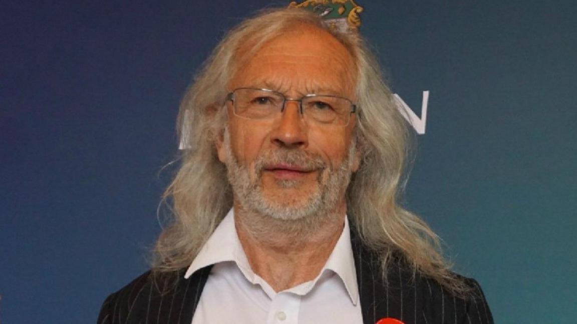 Councillor Phil Riley, who has shoulder length grey hair and is wearing a pinstripe suit jacket over an open-collared white shirt and a Labour Party rosette. 