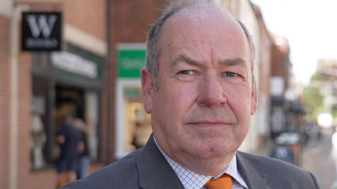 Adrian Owens with grey receding hair wearing a grey suit with an orange tie and white and blue checked shirt. He is standing in a town centre with shops and shoppers behind him.