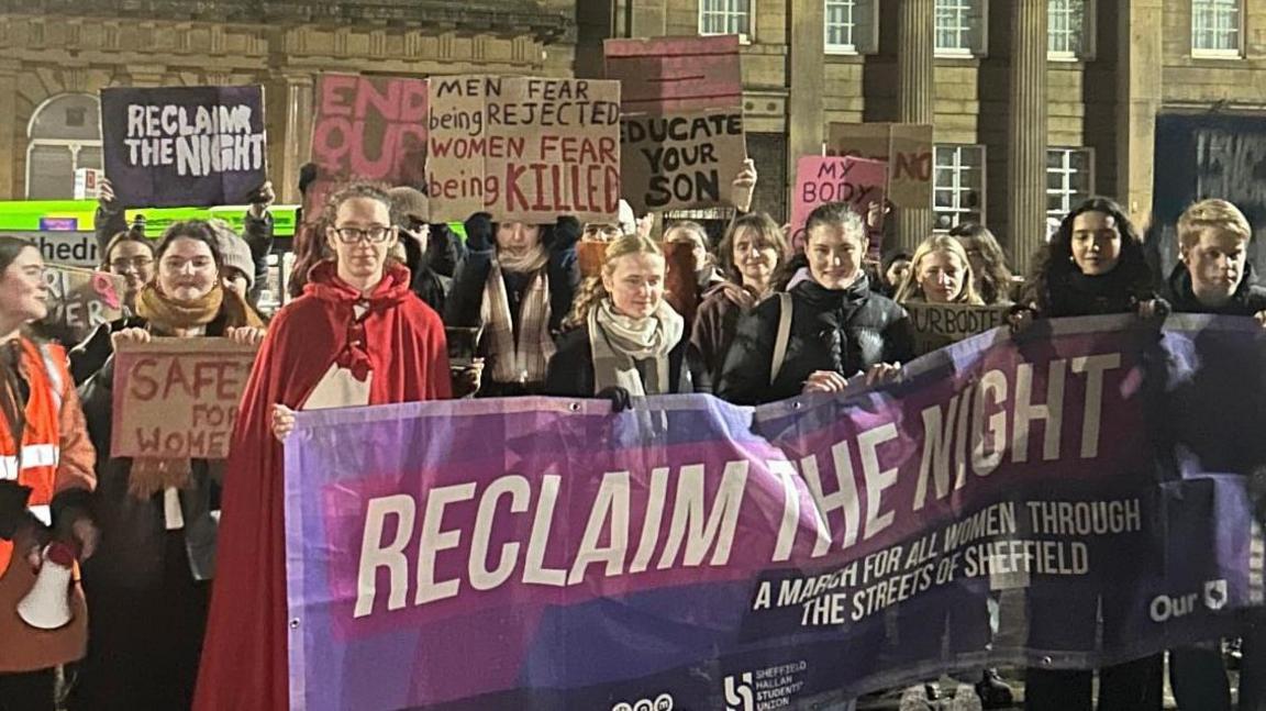 A group of women hold banners in Sheffield city centre. At the front they hold a purple banner reading: Reclaim the night, a march for women through the streets of Sheffield. 