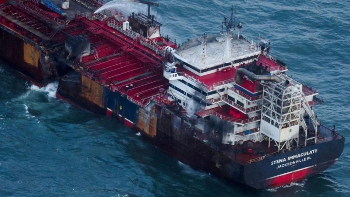 A close up shot of the Stena Immaculate oil tanker ship, the exterior is navy blue and the interior is red. There is water shooting out of the damaged hull.