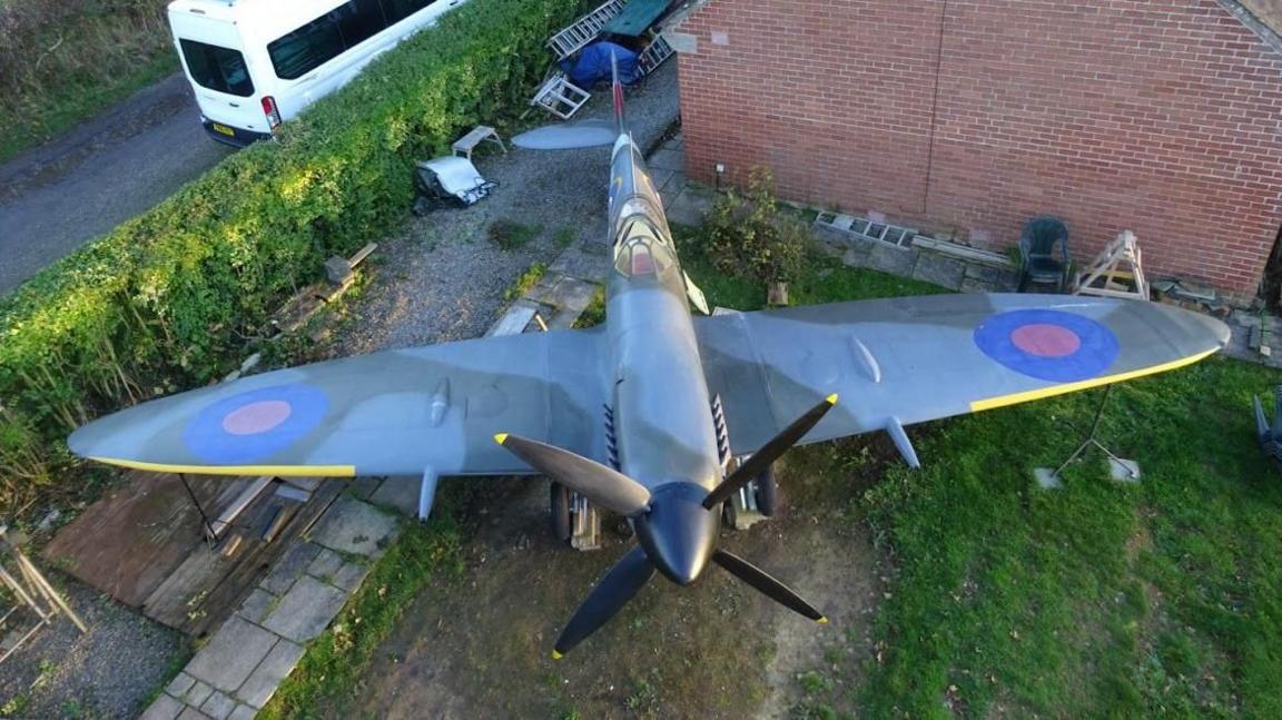 An aerial image of the spitfire in Mr Price's garden. The aircraft is next to the house, a red-brick building. It has the typical colours and markings of a Spitfire, including yellow details on the wings and blades, and red and blue circle on the wings.